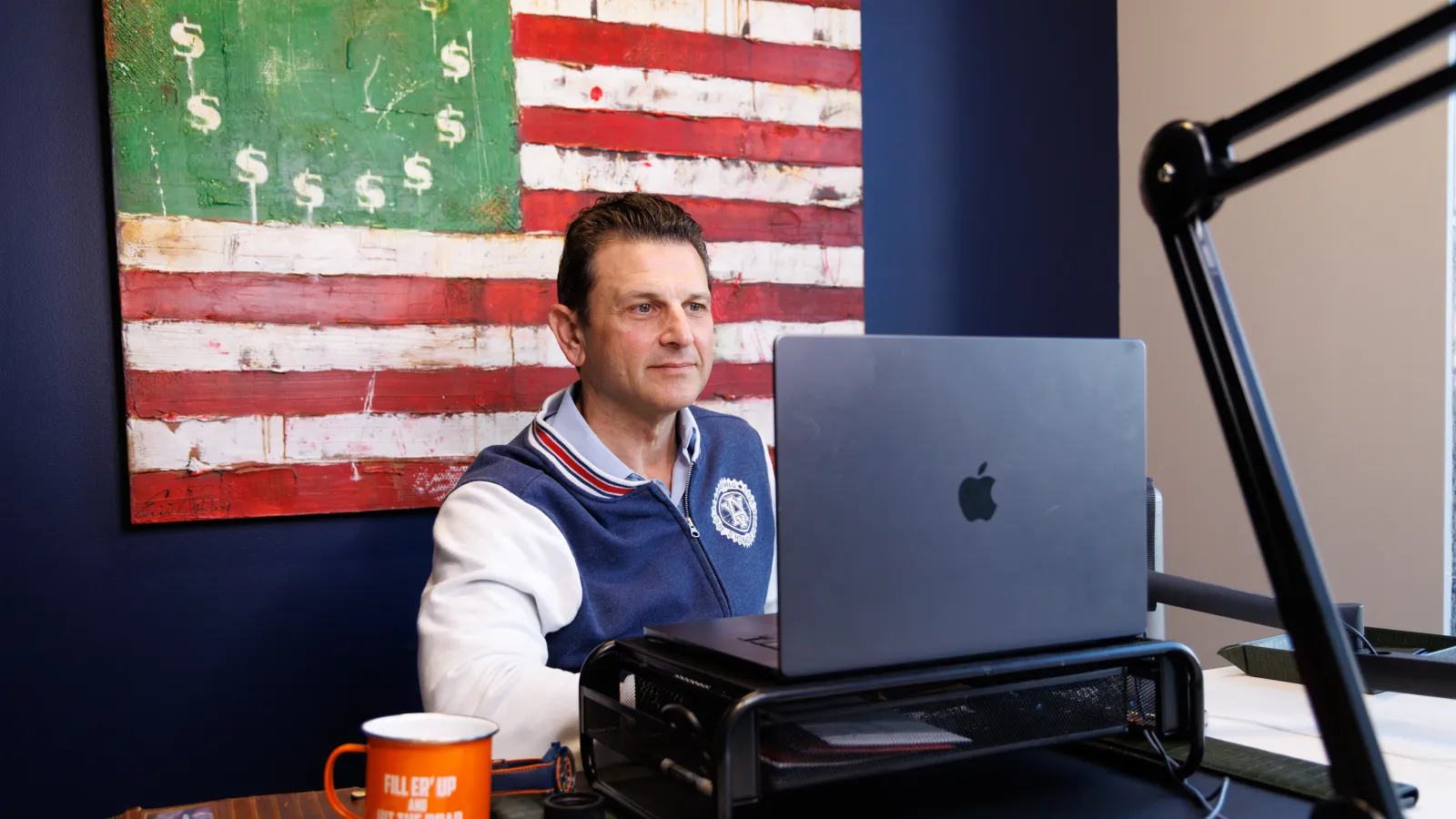 Man working on a laptop at a desk with an American flag painting featuring dollar signs in the background.