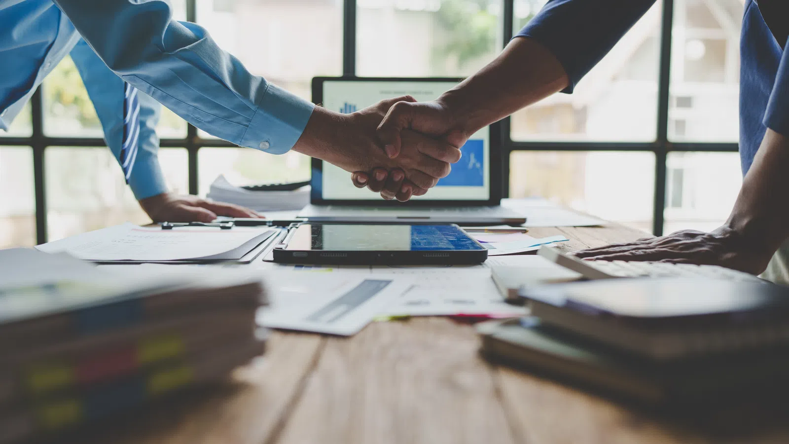 Two Essayon professionals shaking hands over a desk filled with documents and a tablet in a bright office setting