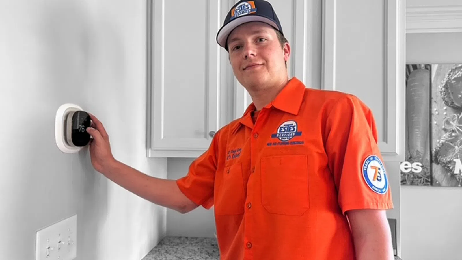 Technician in orange uniform adjusting a smart thermostat on a white wall in a modern kitchen.