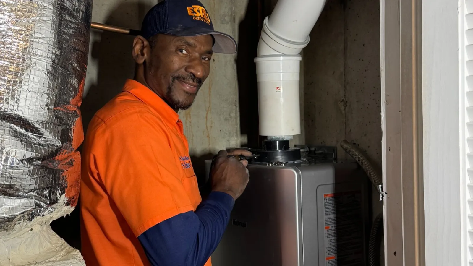 Estes Plumber in orange shirt uniform installing a home water heater in a basement with visible pipes and ducts.