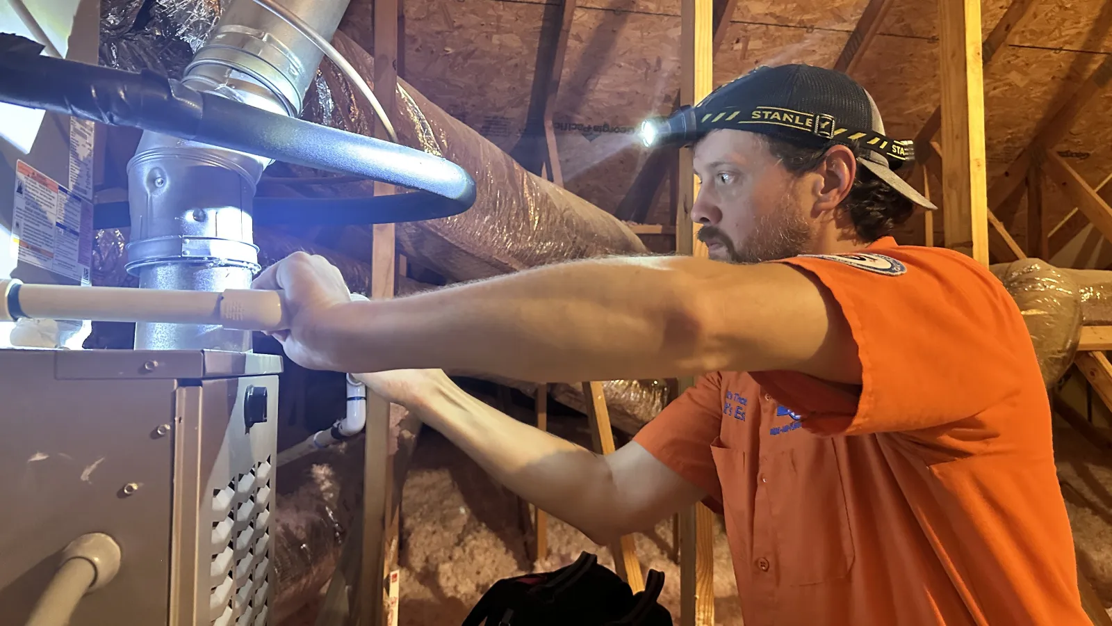 Technician in orange shirt installs HVAC ductwork in attic using headlamp for illumination.