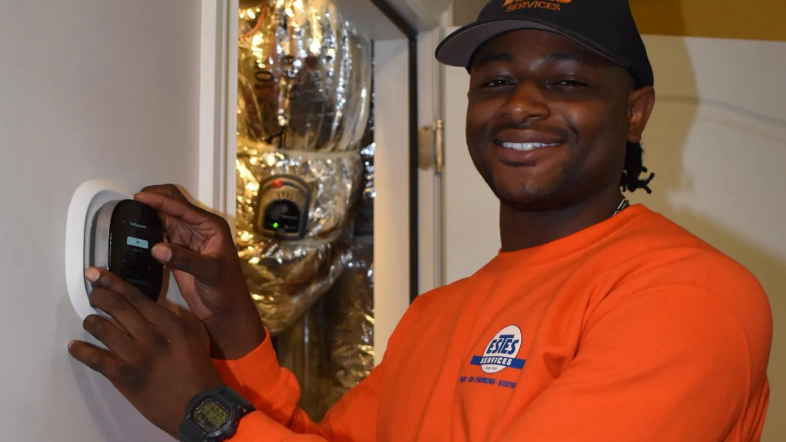 Estes Services Technician in orange shirt installing or inspecting a smoke detector on a white ceiling inside a room with HVAC ducts.