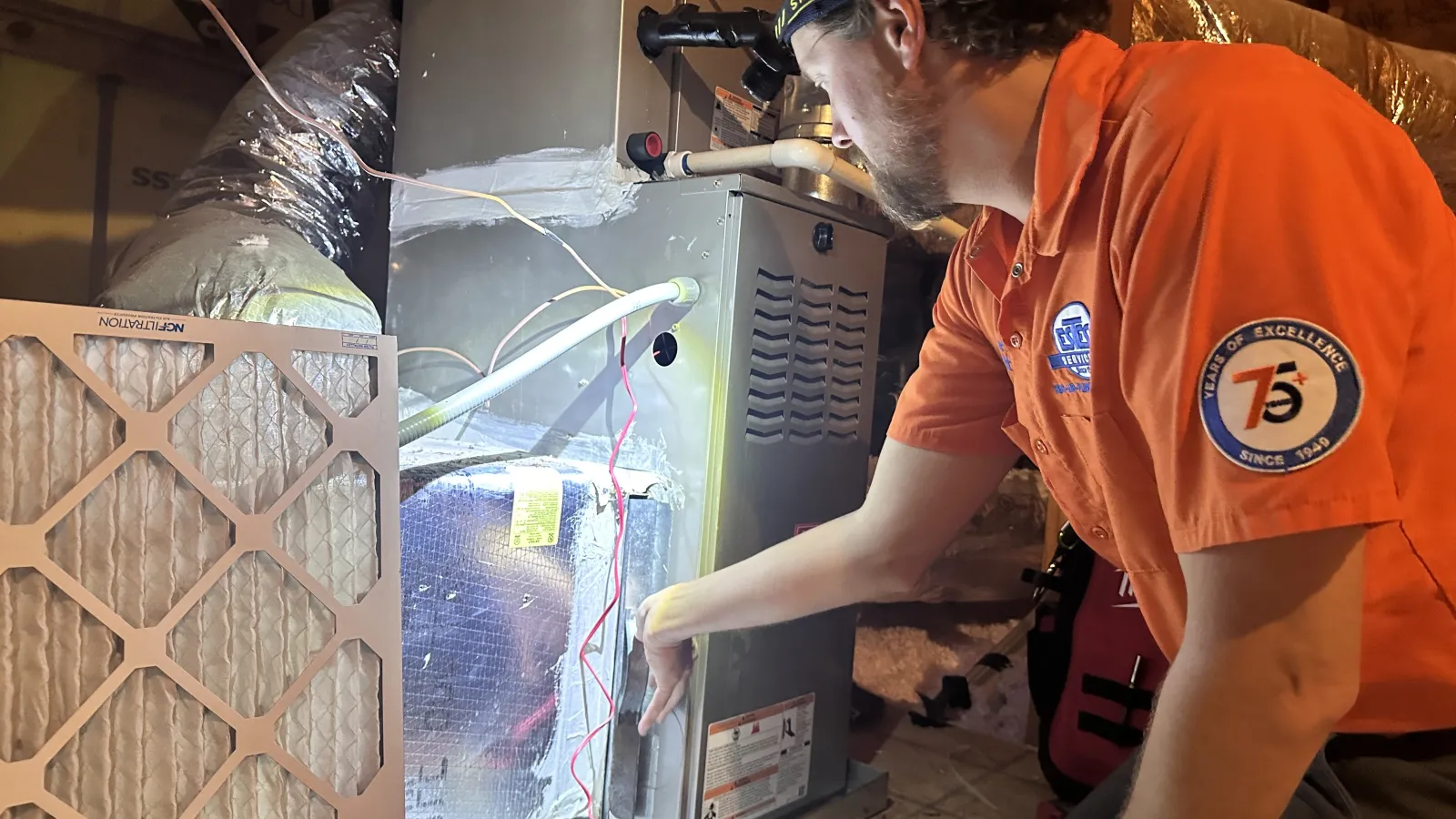 Estes Services Technician in orange shirt inspecting and repairing a furnace system in an attic space with filters and ducts.
