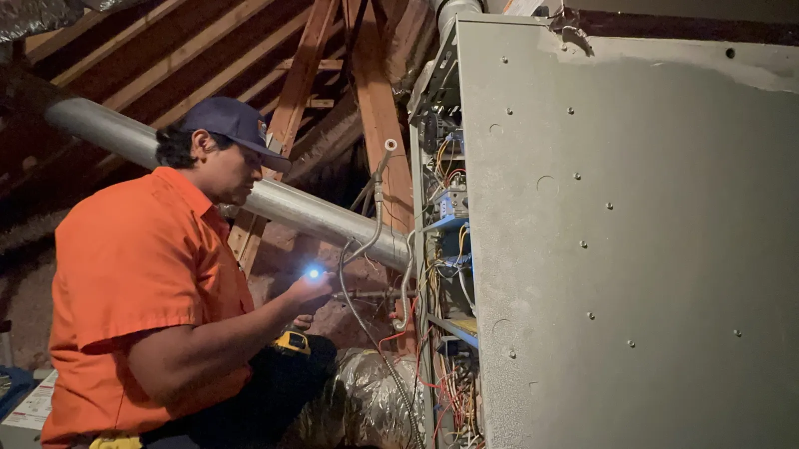 Estes Services Technician in orange uniform furnace repair system in attic with flashlight and tools.