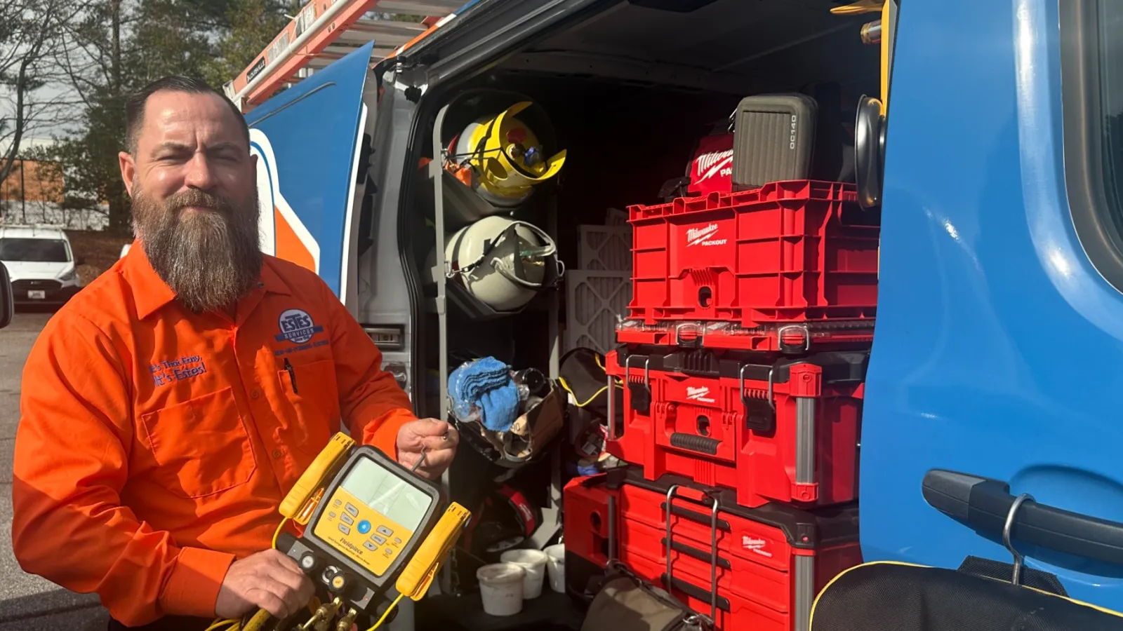 Estes Services Technician in orange uniform holding HVAC diagnostic device beside open service van with equipment and tools.