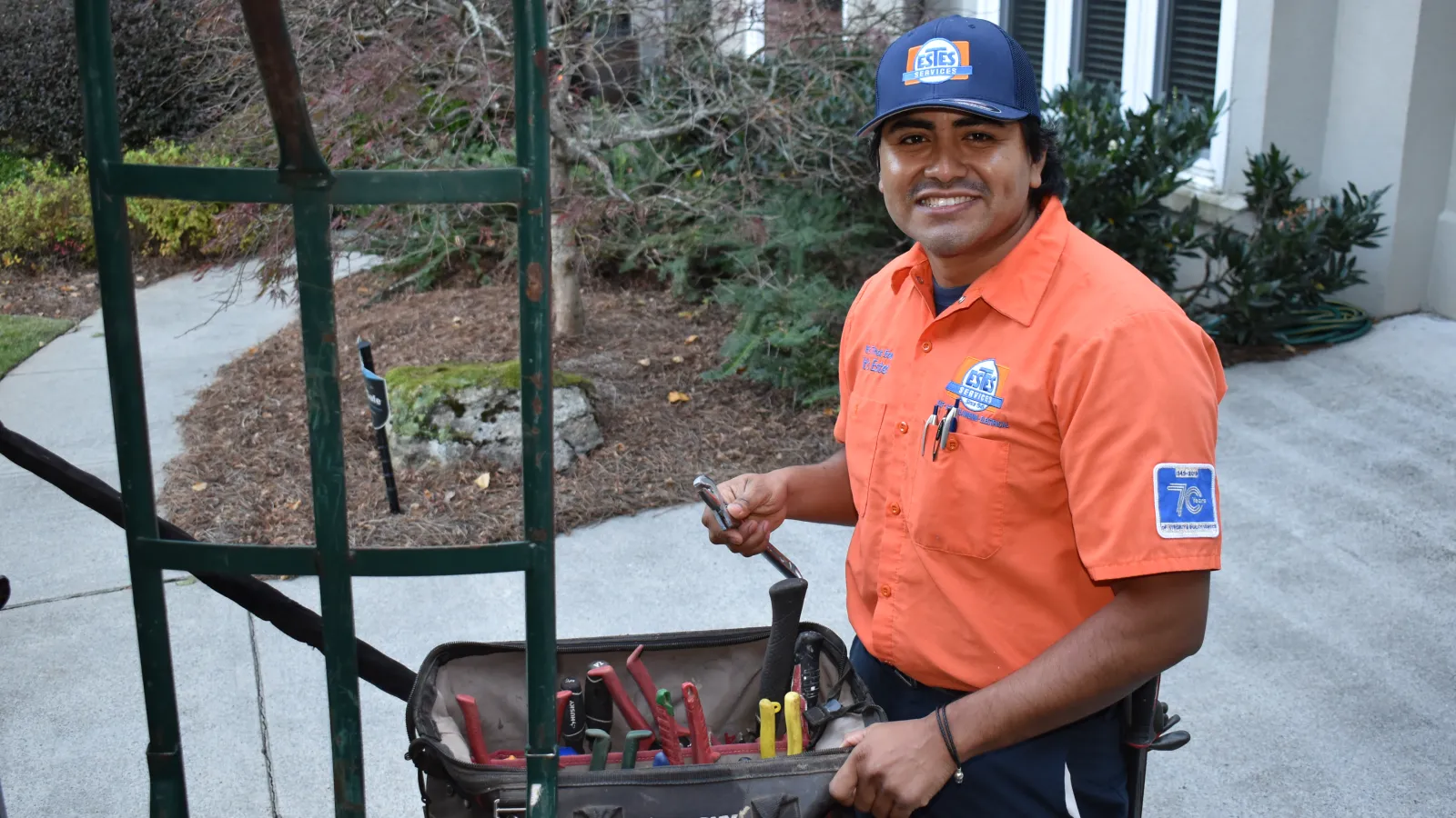 Smiling Estes Services technician in an orange shirt holds a wrench next to a tool bag filled with various tools outside a home.