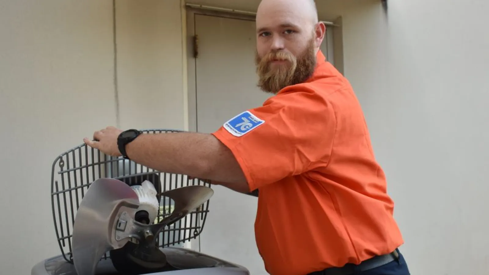 Estes Services Near You. Technician in orange shirt replacing or repairing HVAC unit fan blade outdoors near building.