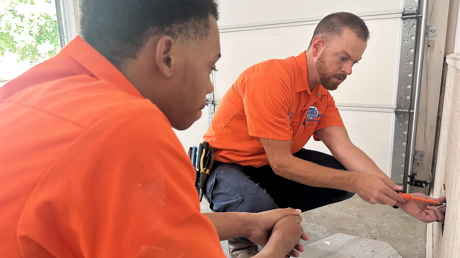 Two Estes Electricians  in orange uniforms repairing an outlet that is not working  inside a garage using hand tools during daytime.