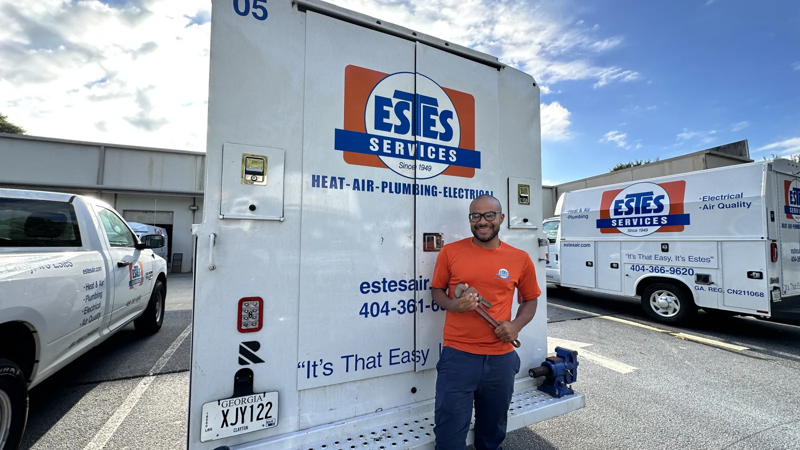 Technician standing by Estes Services work truck holding a pipe wrench in a parking lot under blue sky.