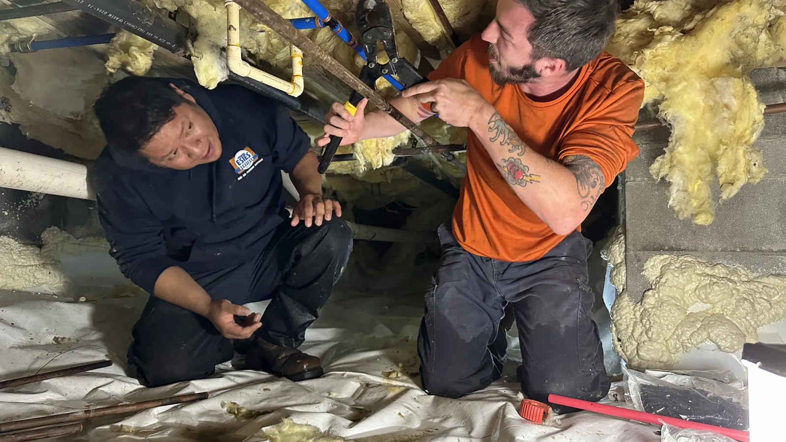 Two Estes plumbers working on pipes inside a crawl space with insulation and tools scattered around