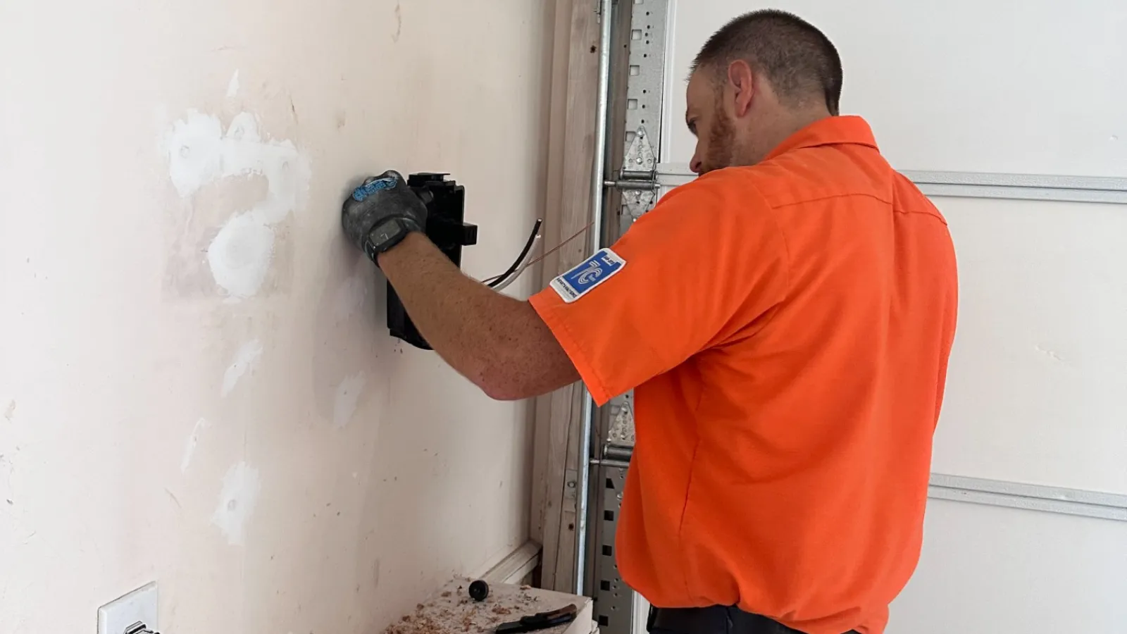 Estes Services Electrician in orange shirt installing or repairing a device on a white wall inside a garage.