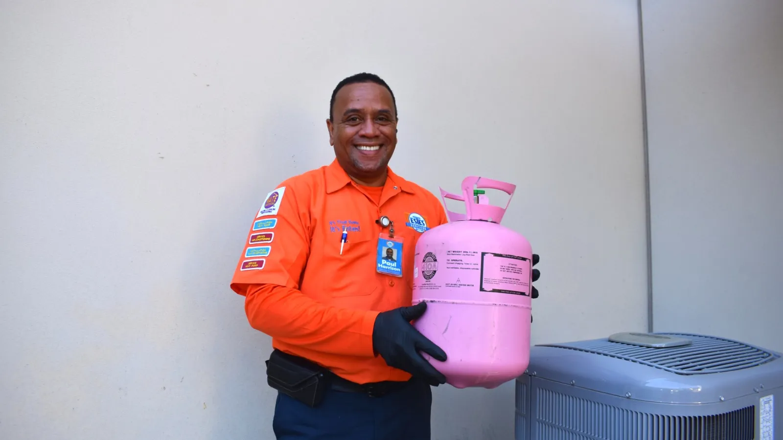 Smiling Estes HVAC technician in uniform holding a refrigerant tank next to an air conditioning unit outdoors.
