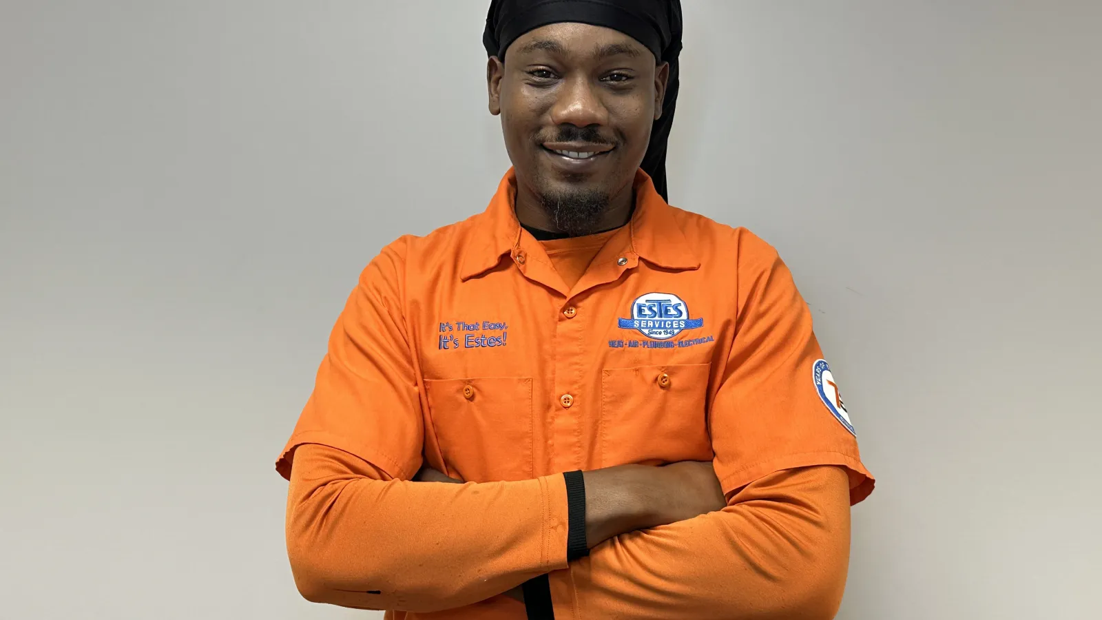 Smiling  Estes Tech in an orange Estes Services uniform with arms crossed against a plain background.