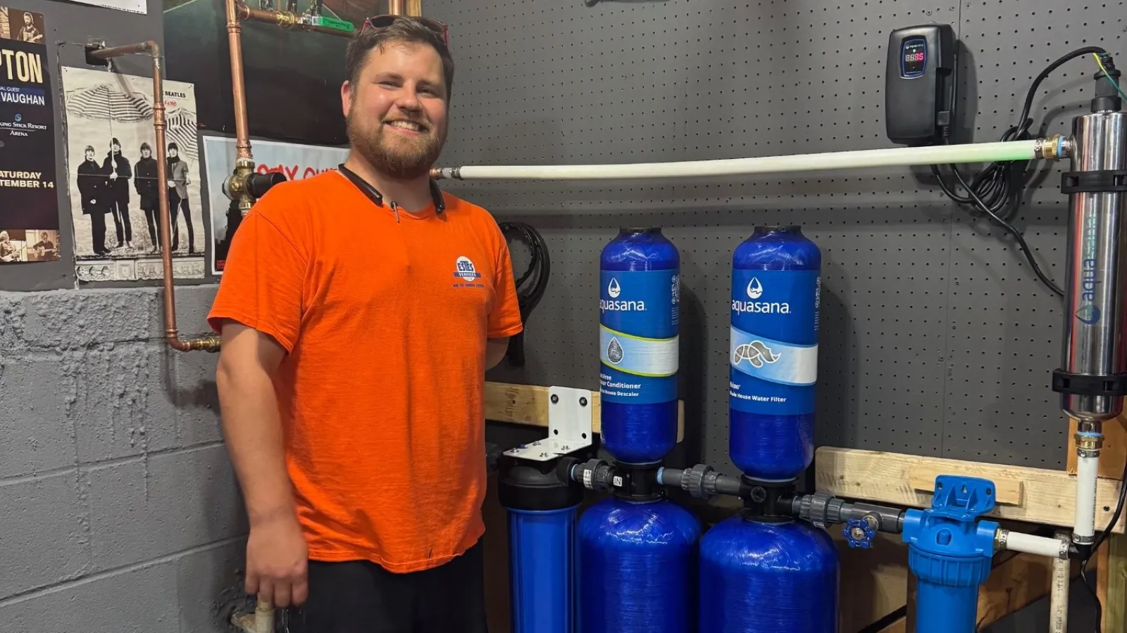 Estes Plumber in an orange uniform stands next to a water filtration system with blue tanks in a workshop setting