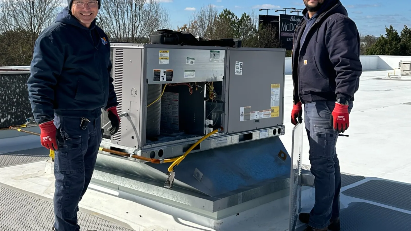 Two Estes Commercial technicians in winter clothing working on rooftop HVAC unit under clear blue sky.