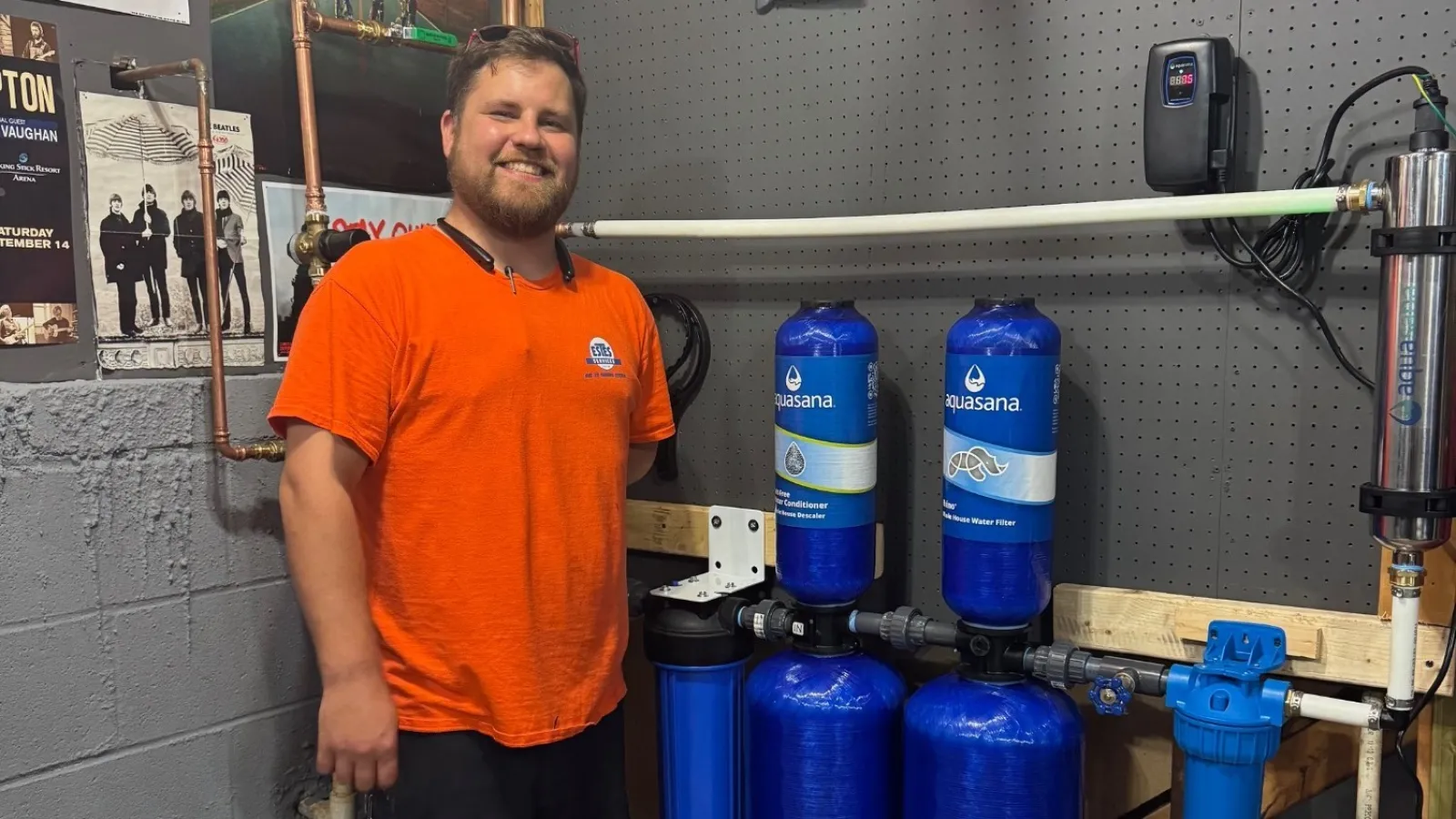 Estes Plumber in orange shirt stands next to large blue water filtration system in workshop with tools and posters.