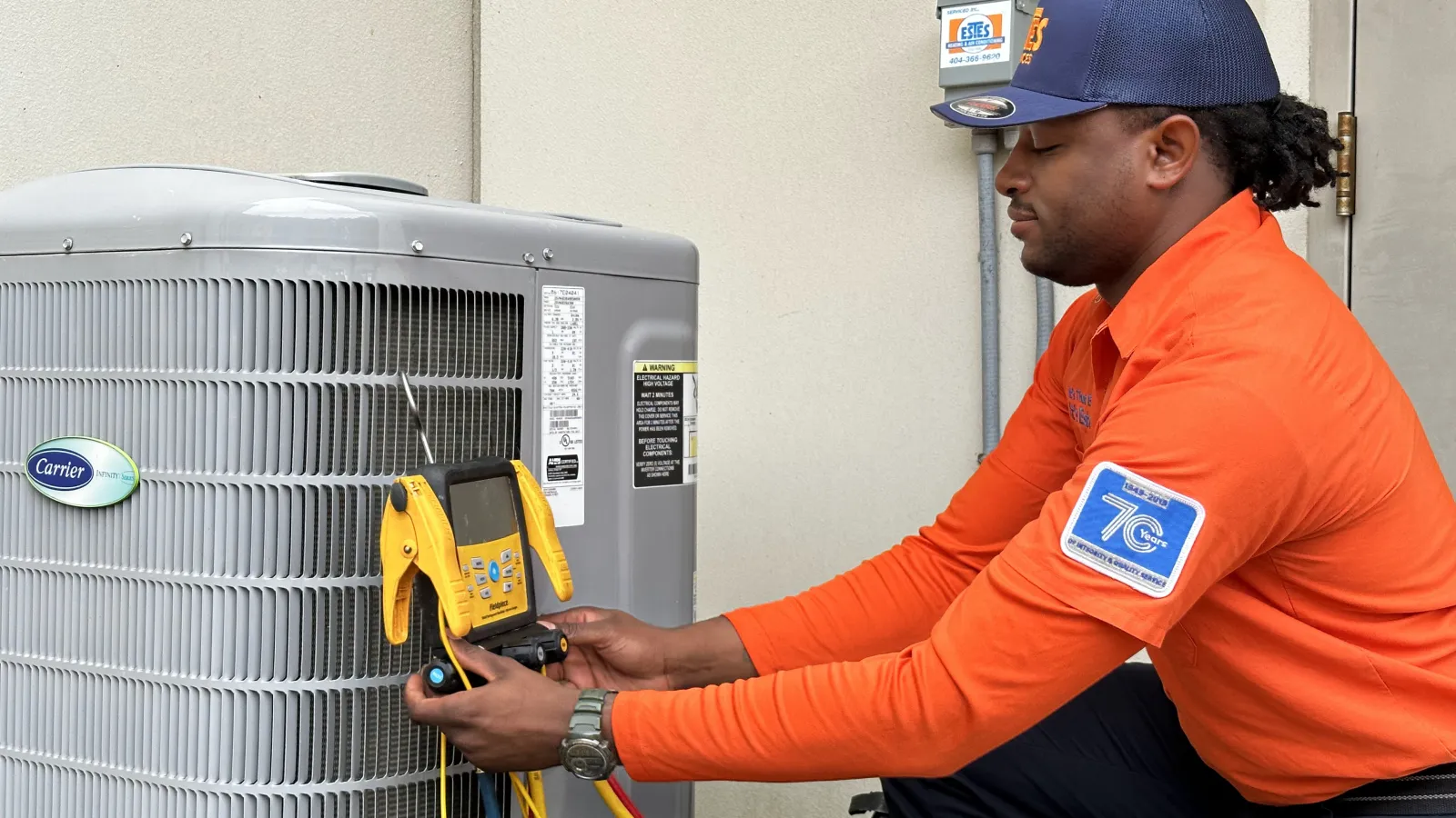 Estes Services Heat Pump Technician in orange uniform testing a Carrier HVAC unit with diagnostic equipment outdoors.