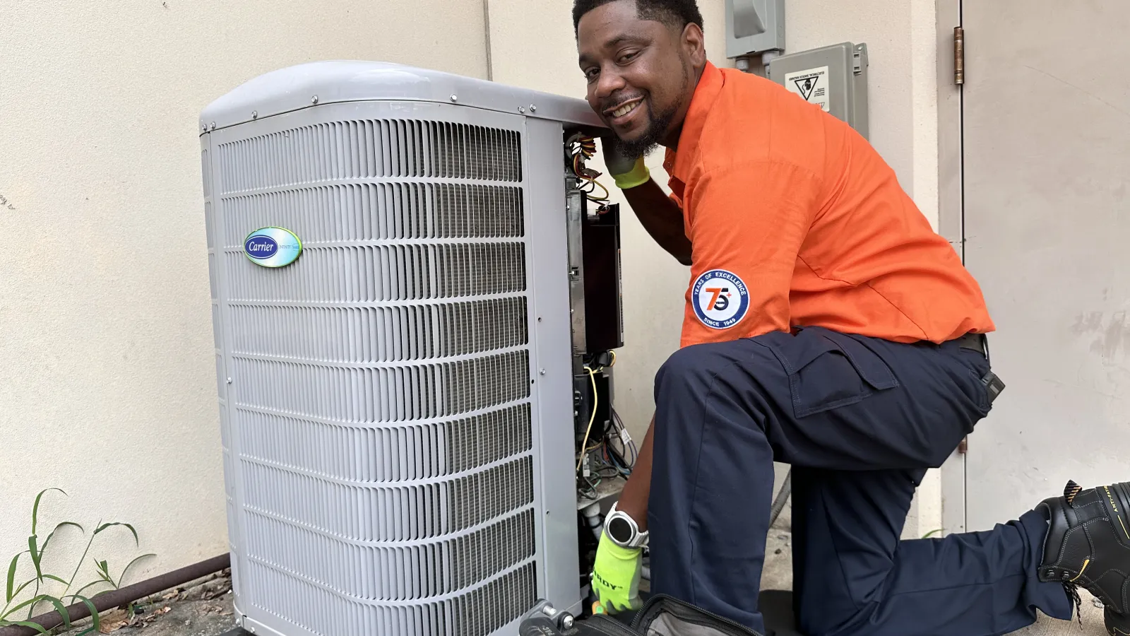 Estes Heating and Air Technician in orange shirt uniform working on outdoor Carrier air conditioning unit with tools on ground in building exterior.