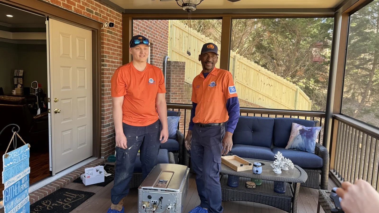 Two plumbers workers in orange shirts standing on a screened porch with a tankless water heater ready to be installed