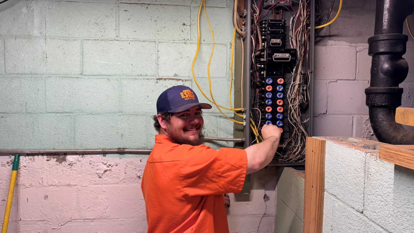 Estes Electrician in a uniform shirt working on a circuit breaker panel in a basement with exposed wires and concrete walls