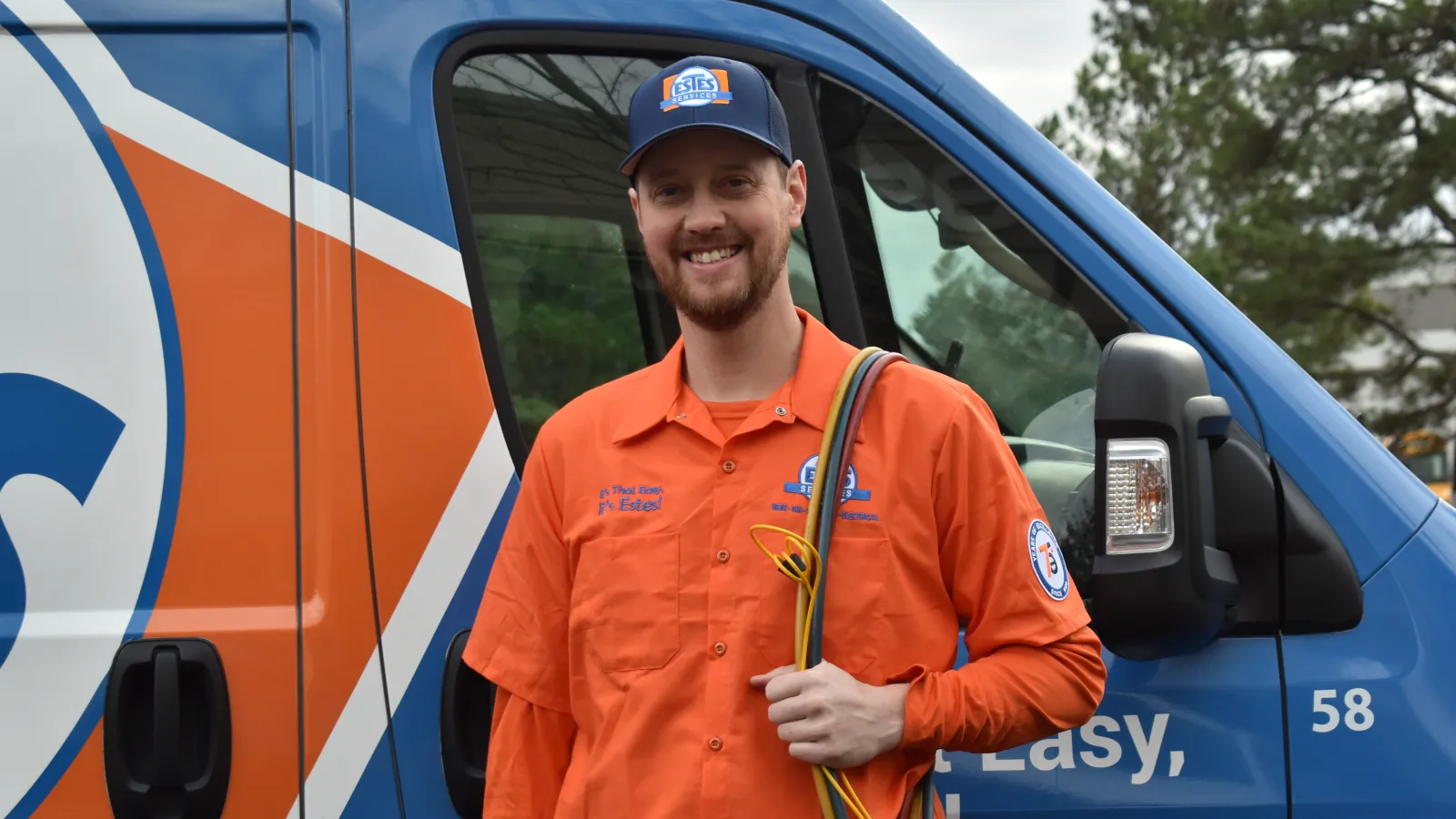 Smiling electrician in orange uniform holding wires stands next to branded blue and orange service van outdoors.