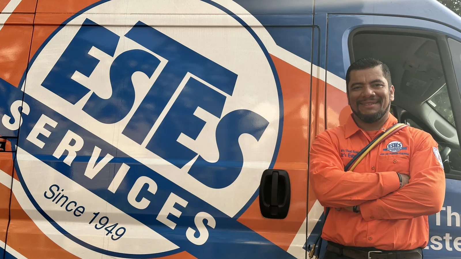 Smiling Estes HVAC technician in orange uniform stands with arms crossed next to Estes Services company van logo.