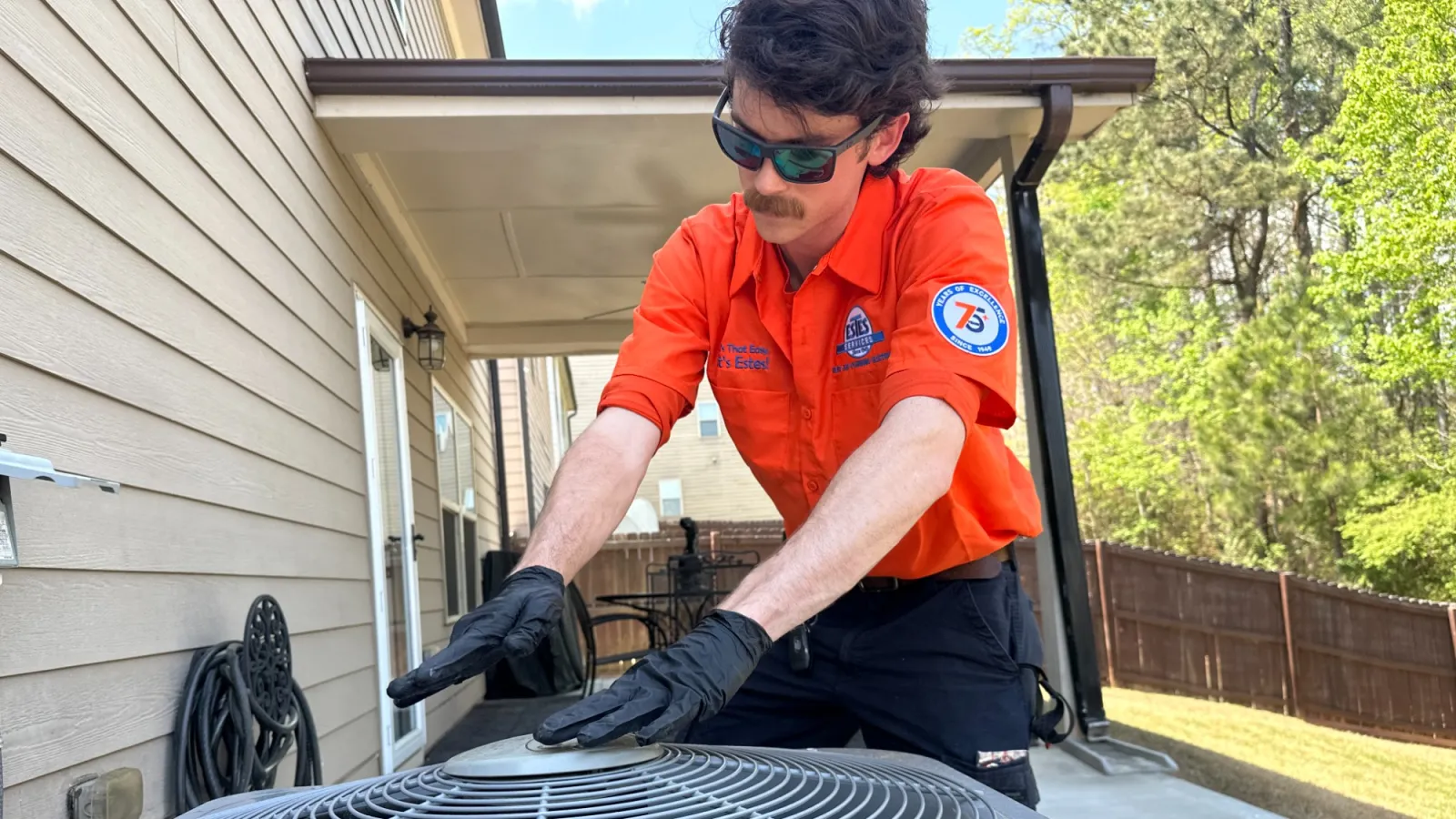 Estes Services HVAC technician in orange shirt inspecting outdoor air conditioning unit on a sunny day.