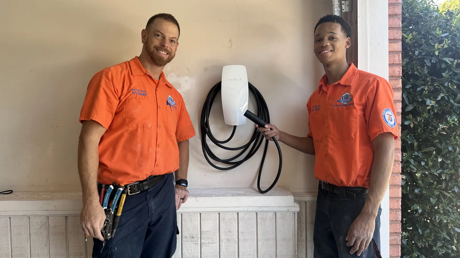 Two licensed electricians in Estes Services uniform  shirts installing or demonstrating a Tesla electric vehicle charger in a garage space.