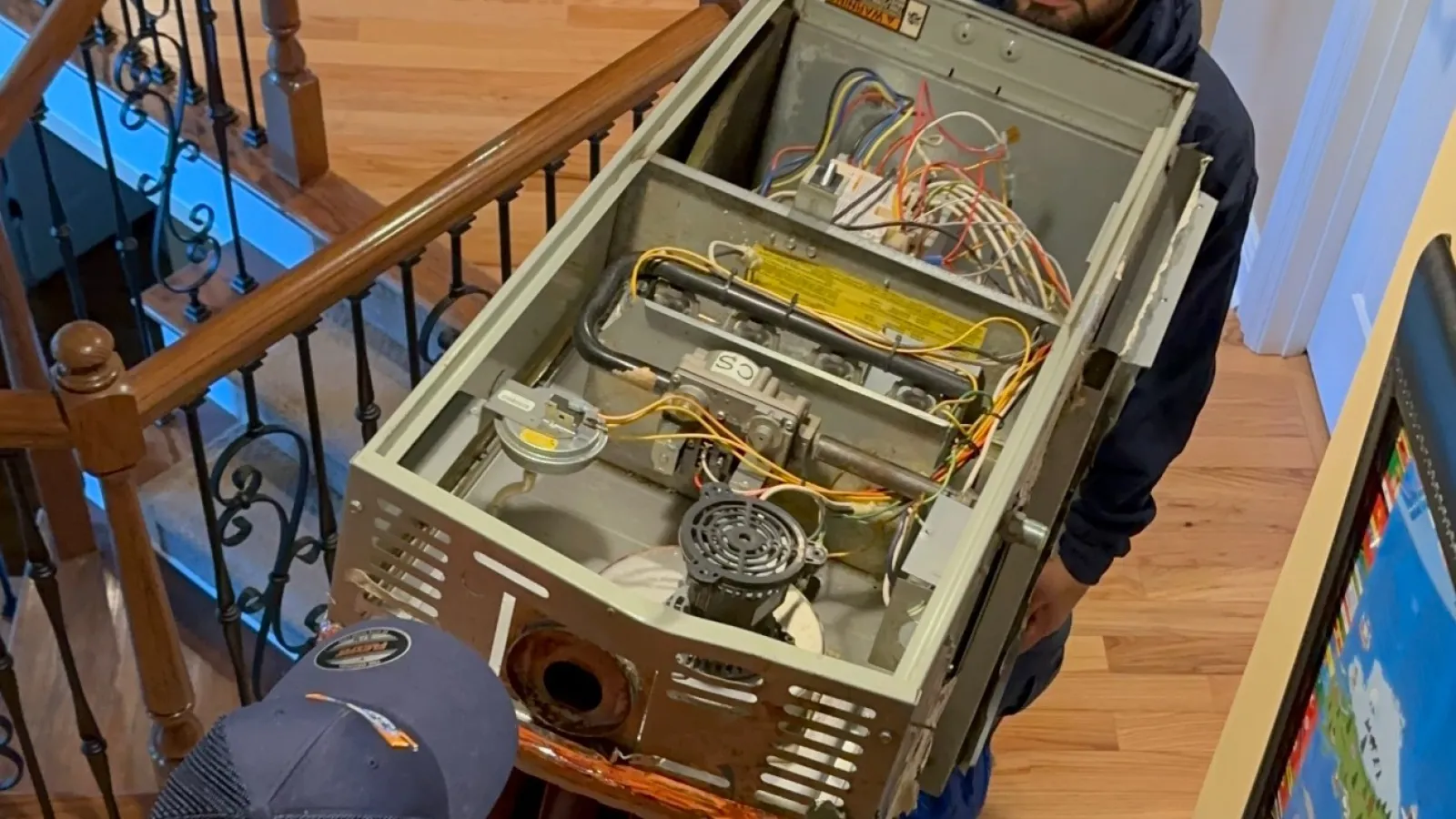 Two Estes Installers workers carrying a large Carrier Furnace unit up wooden stairs inside a home with metal railings and hardwood floors.