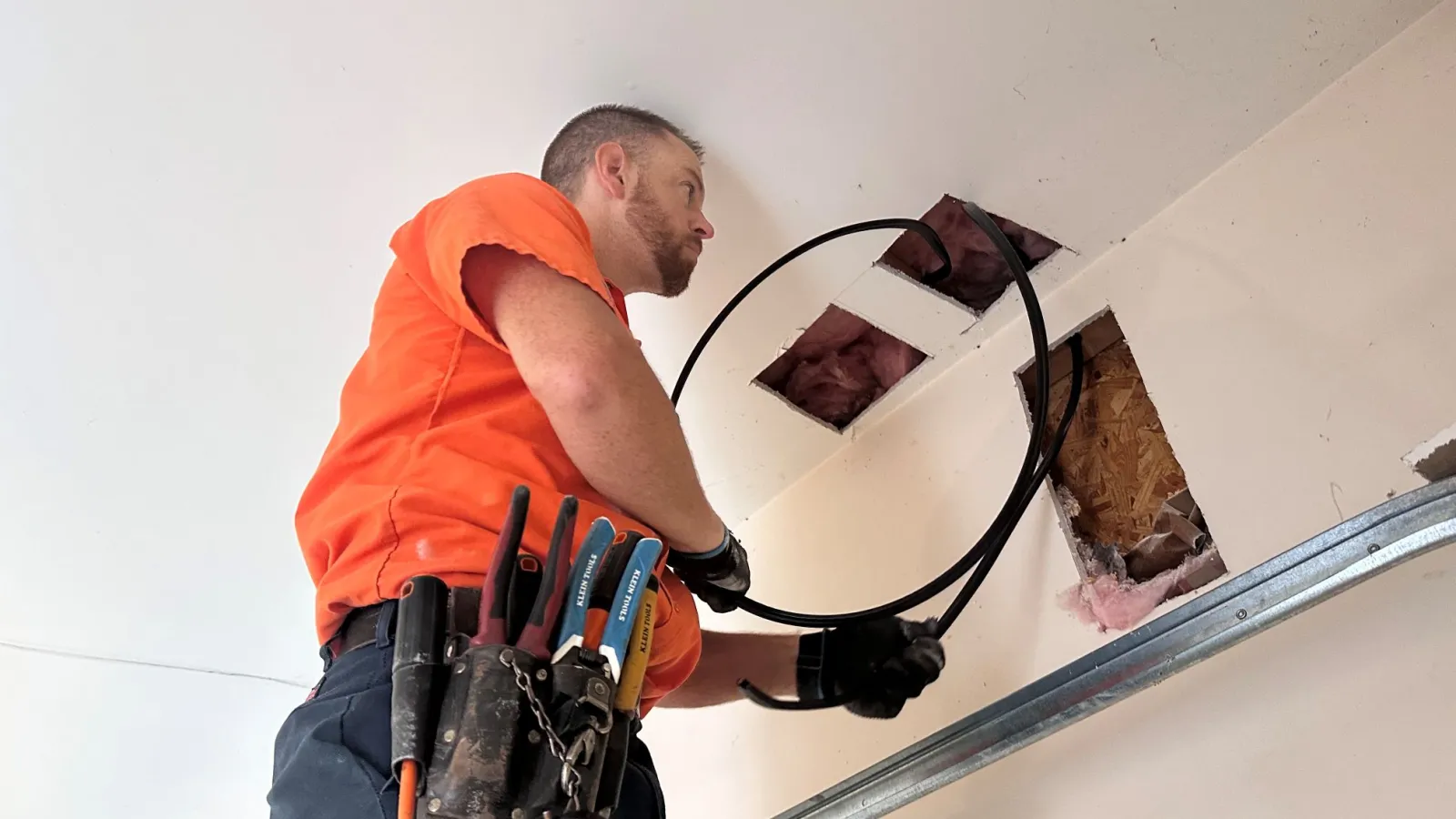 Electrician of Estes Services in orange shirt installing wiring through ceiling with tools in belt and gloves.