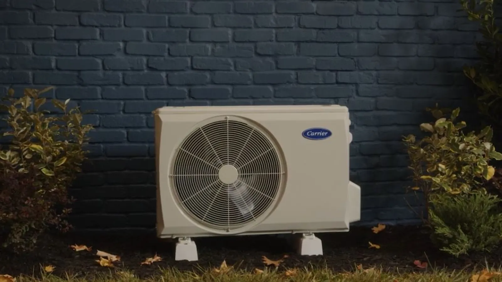 Outdoor Carrier air conditioning unit installed on grass against a dark blue brick wall with surrounding plants.
