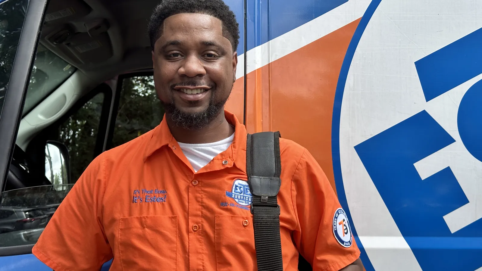 Smiling Darryl, Estes service technician in orange uniform standing next to a branded company van with a shoulder bag strap.
