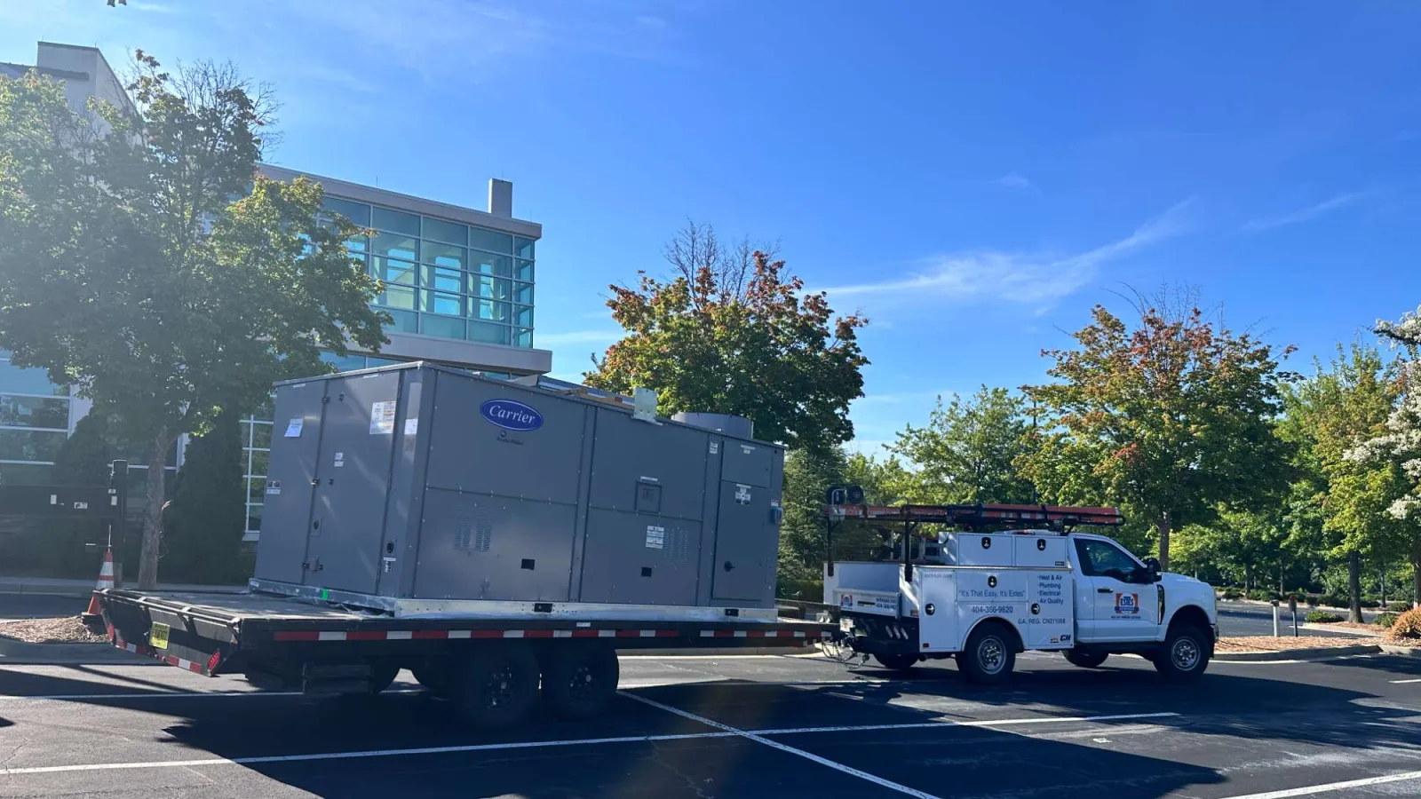 White Estes truck towing flatbed trailer with large Carrier HVAC unit parked in lot near church