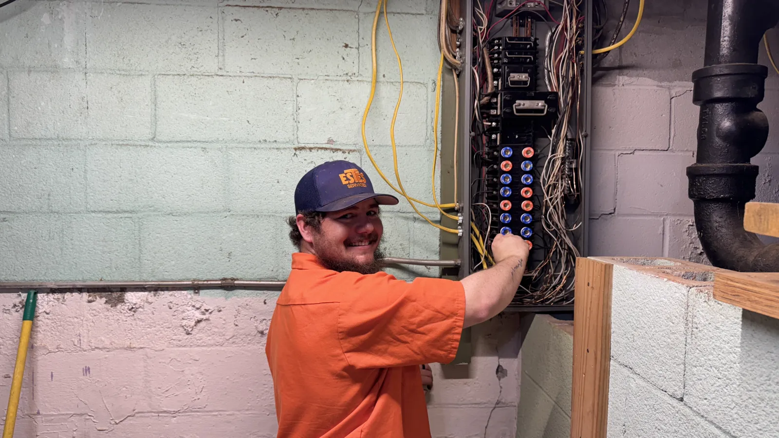 Estes Electrician in a uniform shirt working on a circuit breaker panel in a basement with exposed wires and concrete walls