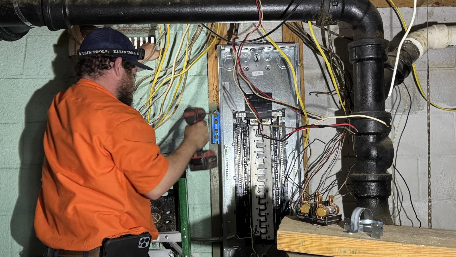 Estes Electrician in orange uniform working on electrical panel with wires and tools in basement setting.