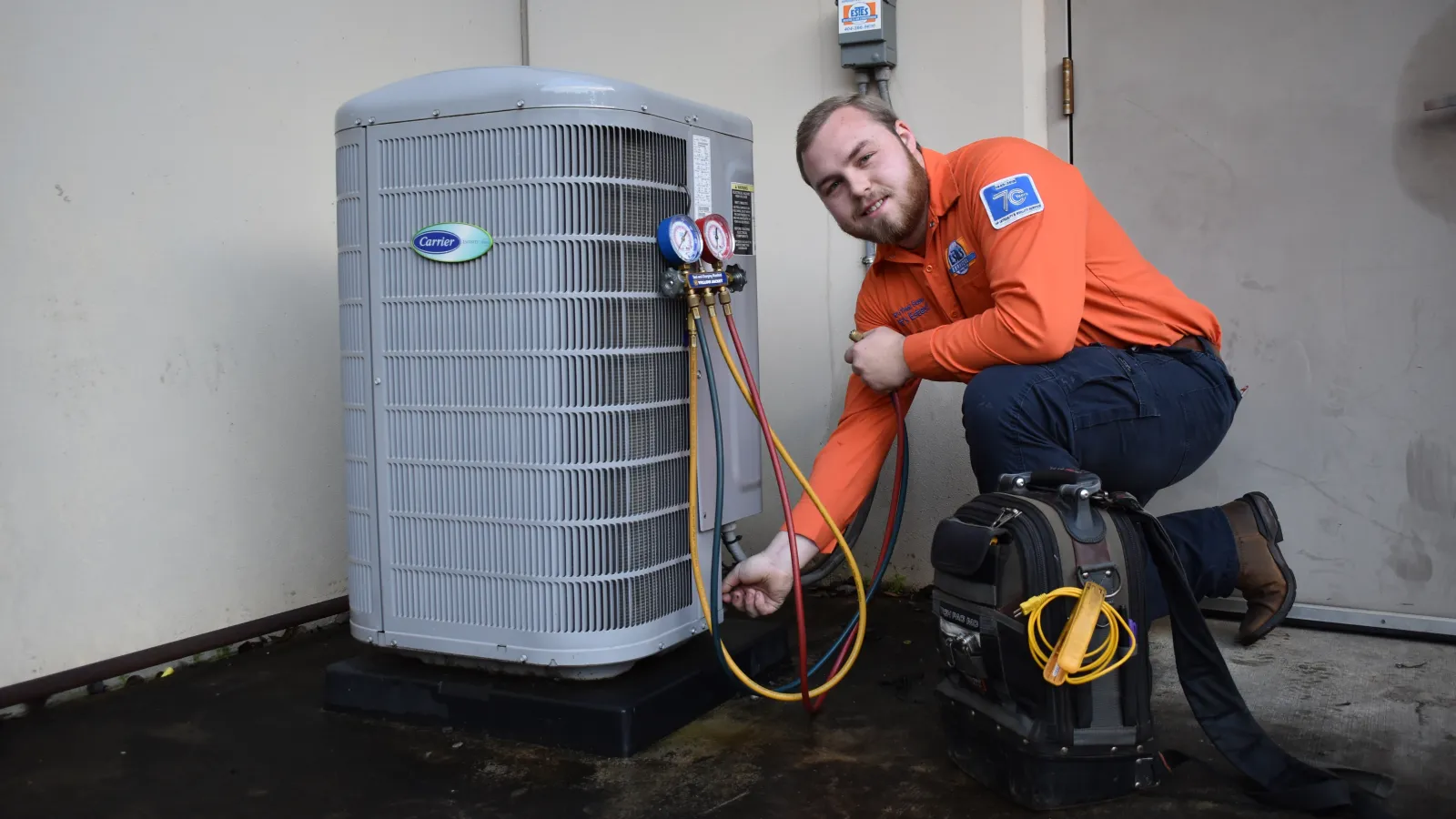 Estes Technician in orange uniform servicing a Carrier HVAC unit with gauges and tools on a black platform.