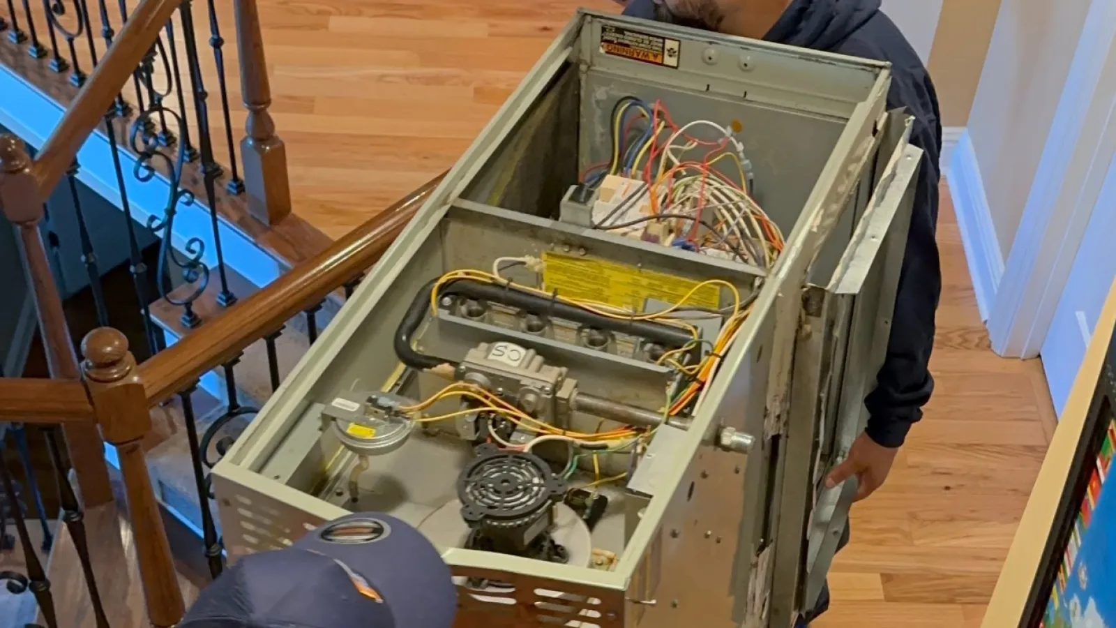 Estes Services men carrying a large Carrier Furnace unit upstairs on a wooden staircase in a home with hardwood floors.