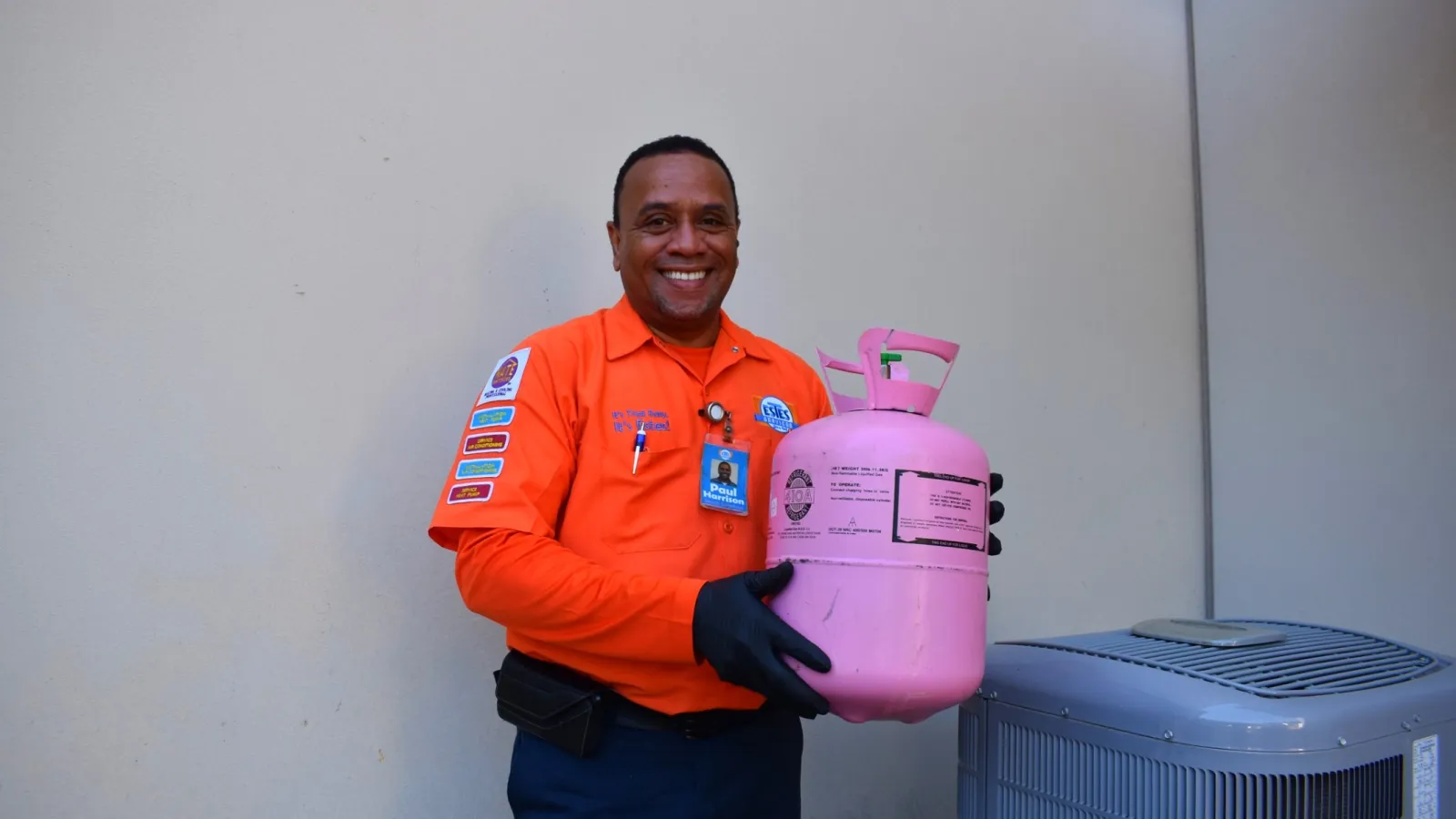 Estes Services Technician in orange shirt uniform holding a pink refrigerant tank next to an air conditioning unit against gray wall.
