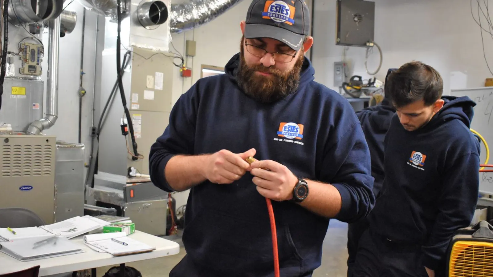 Two Estes Tech Builder Students in Estes  hoodies working with HVAC equipment and tools in a workshop setting.