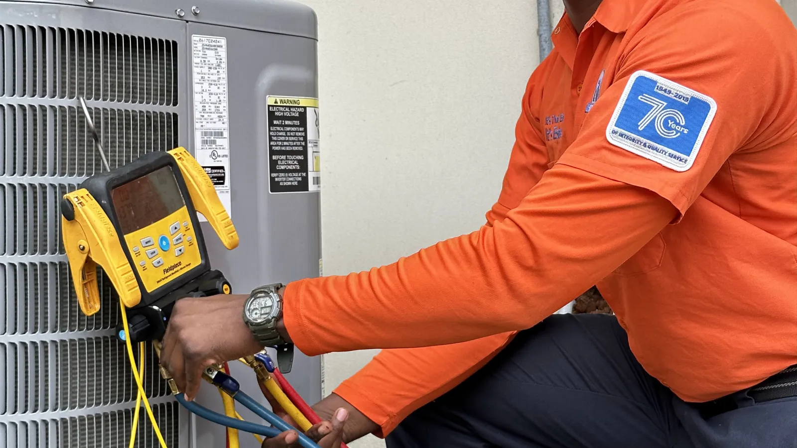 Estes Technician in orange uniform adjusting HVAC unit using digital manifold gauge with red, blue, and yellow hoses.