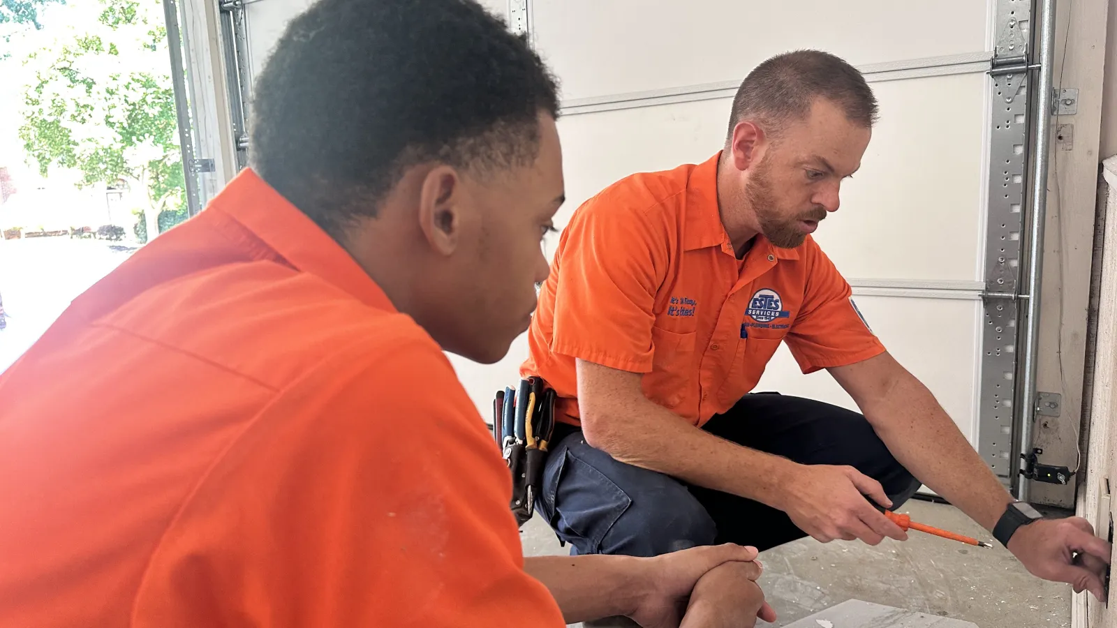 Estes Services Electricians  in orange uniforms repairing power outlet that is not working  inside a garage using hand tools during daytime.