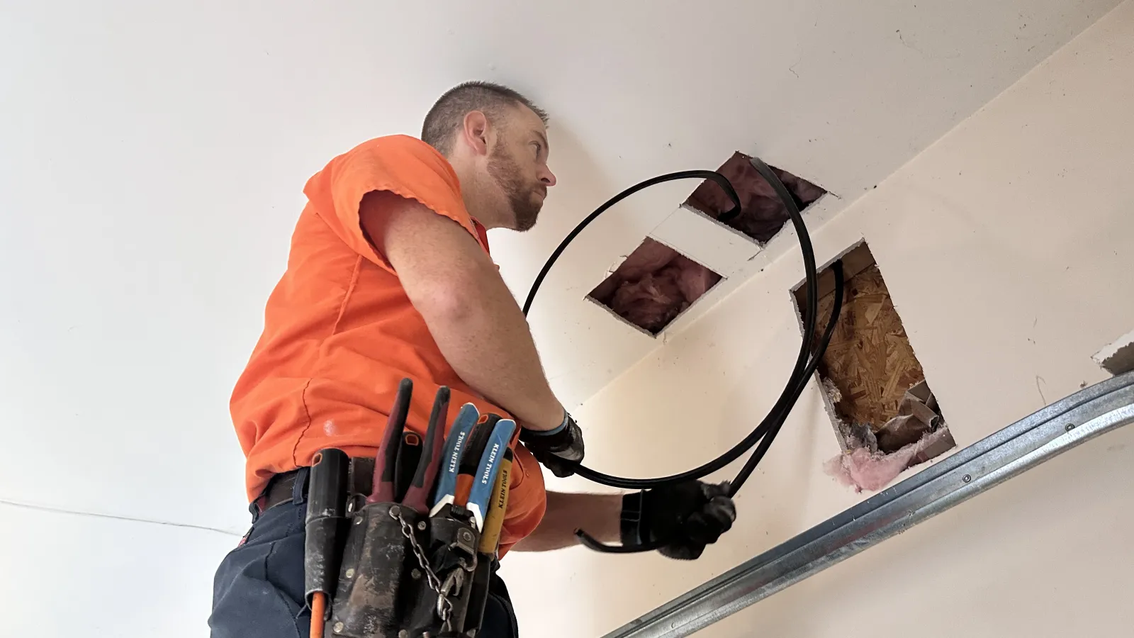 Electrician of Estes Services in orange shirt installing wiring through ceiling with tools in belt and gloves.