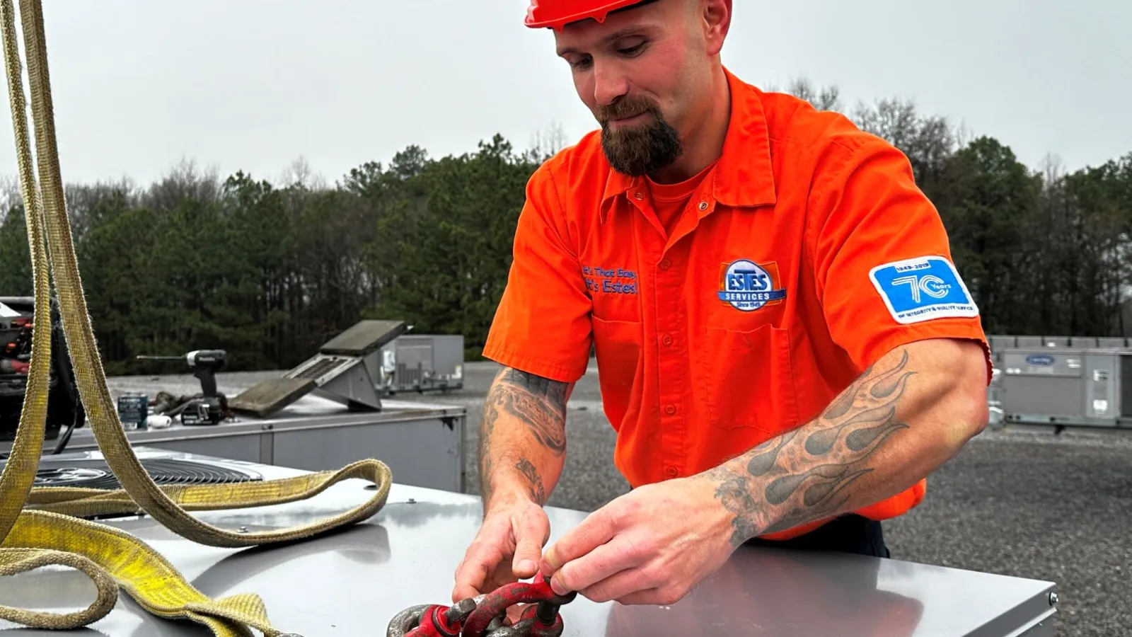 Technician in orange uniform and helmet securing chains on rooftop HVAC unit outdoors on cloudy day