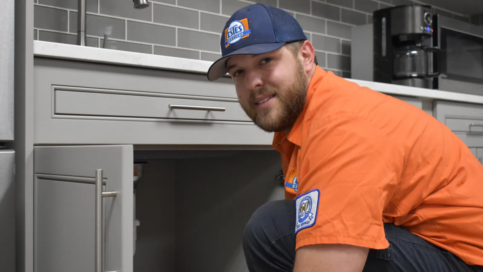 Estes Plumber in orange uniform and cap working under kitchen sink in modern kitchen with gray tiled backsplash.