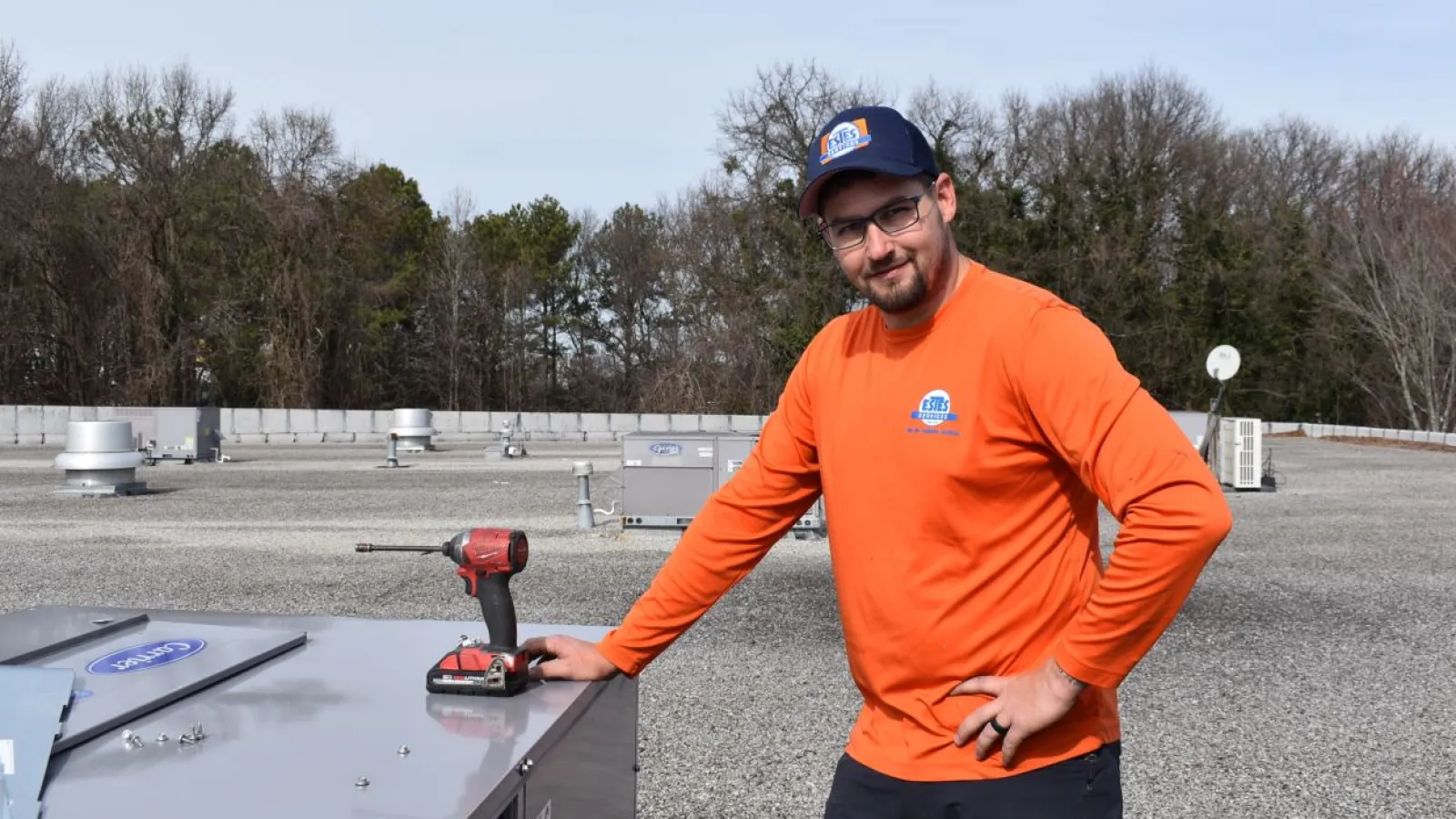 Estes Technician in orange shirt and cap stands on rooftop next to HVAC unit with drill and tool on metal surface.