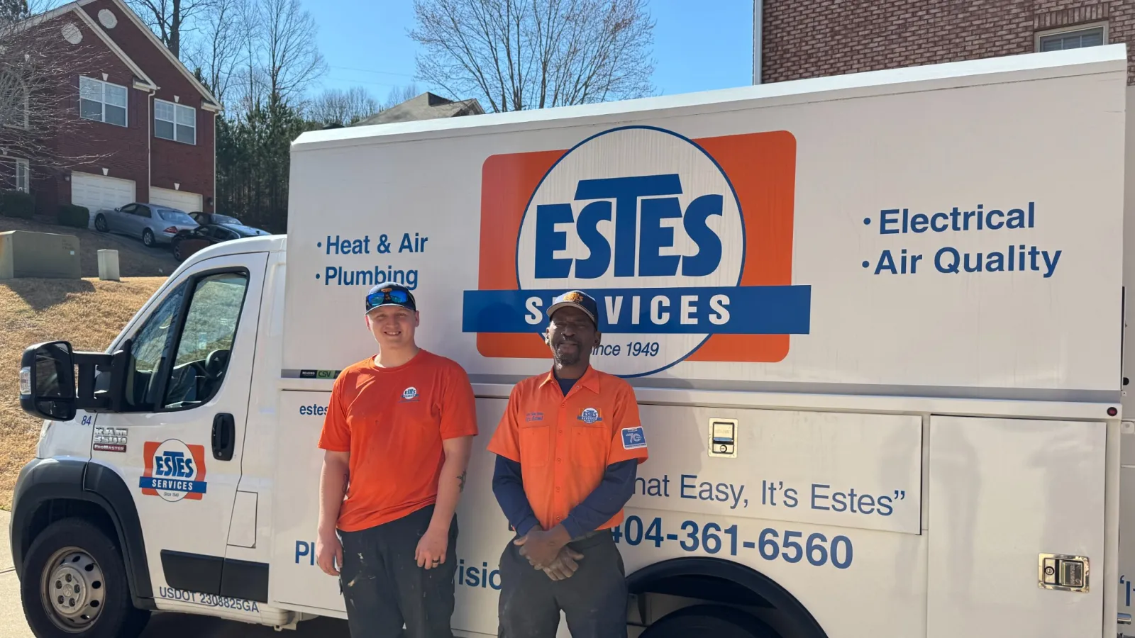 Two Estes Services plumbers standing in front of a branded service van parked outside a residential area on a sunny day