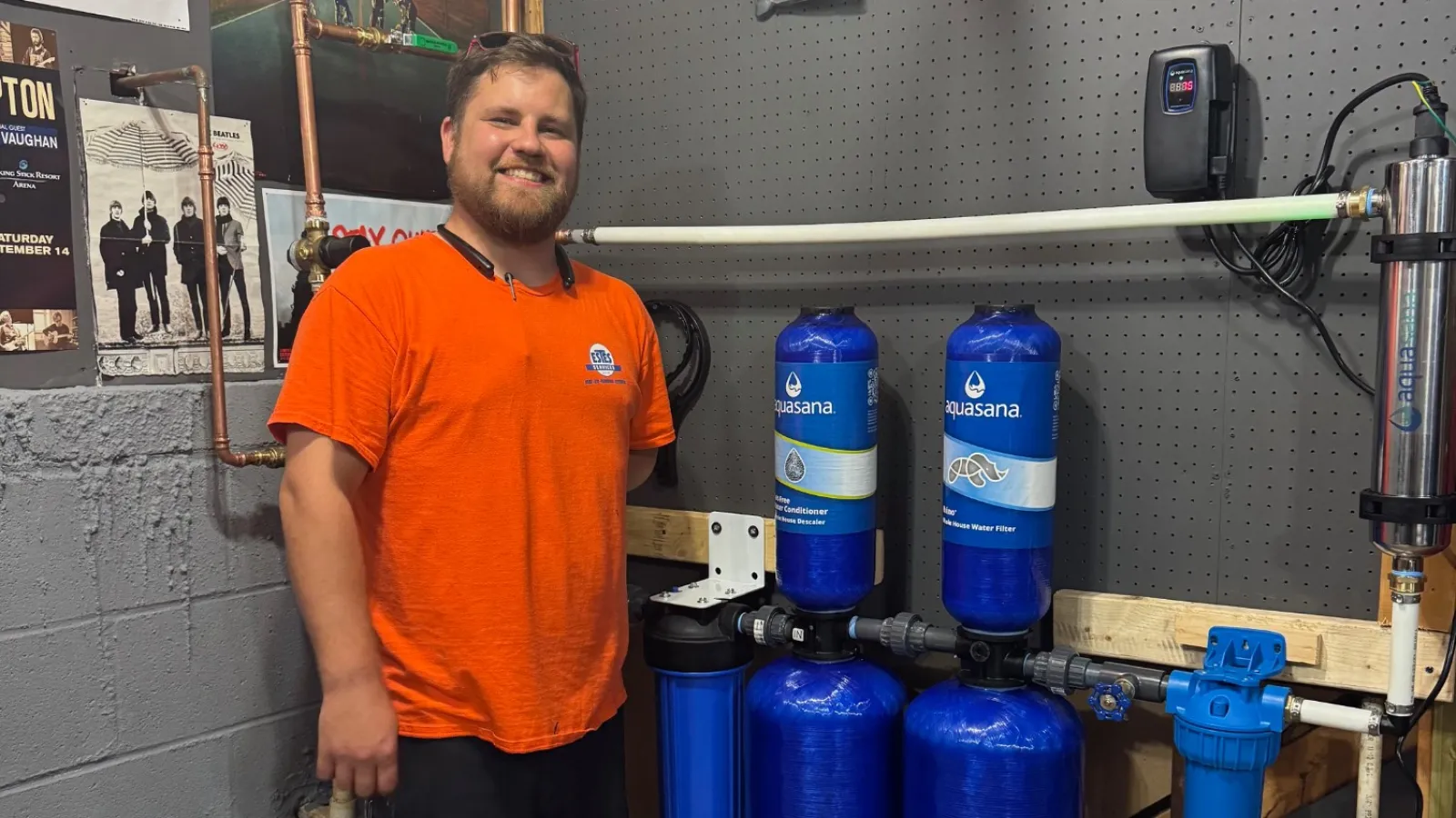 Estes Plumber in orange shirt stands next to large blue water filtration system in workshop with tools and posters.