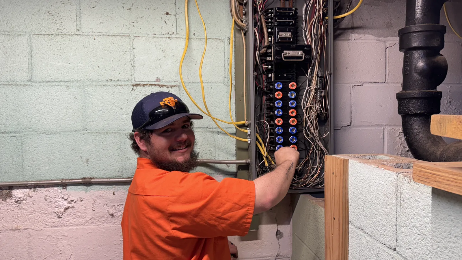 Estes Services Electrician in uniform shirt repairing circuit breaker panel with wiring in basement utility room