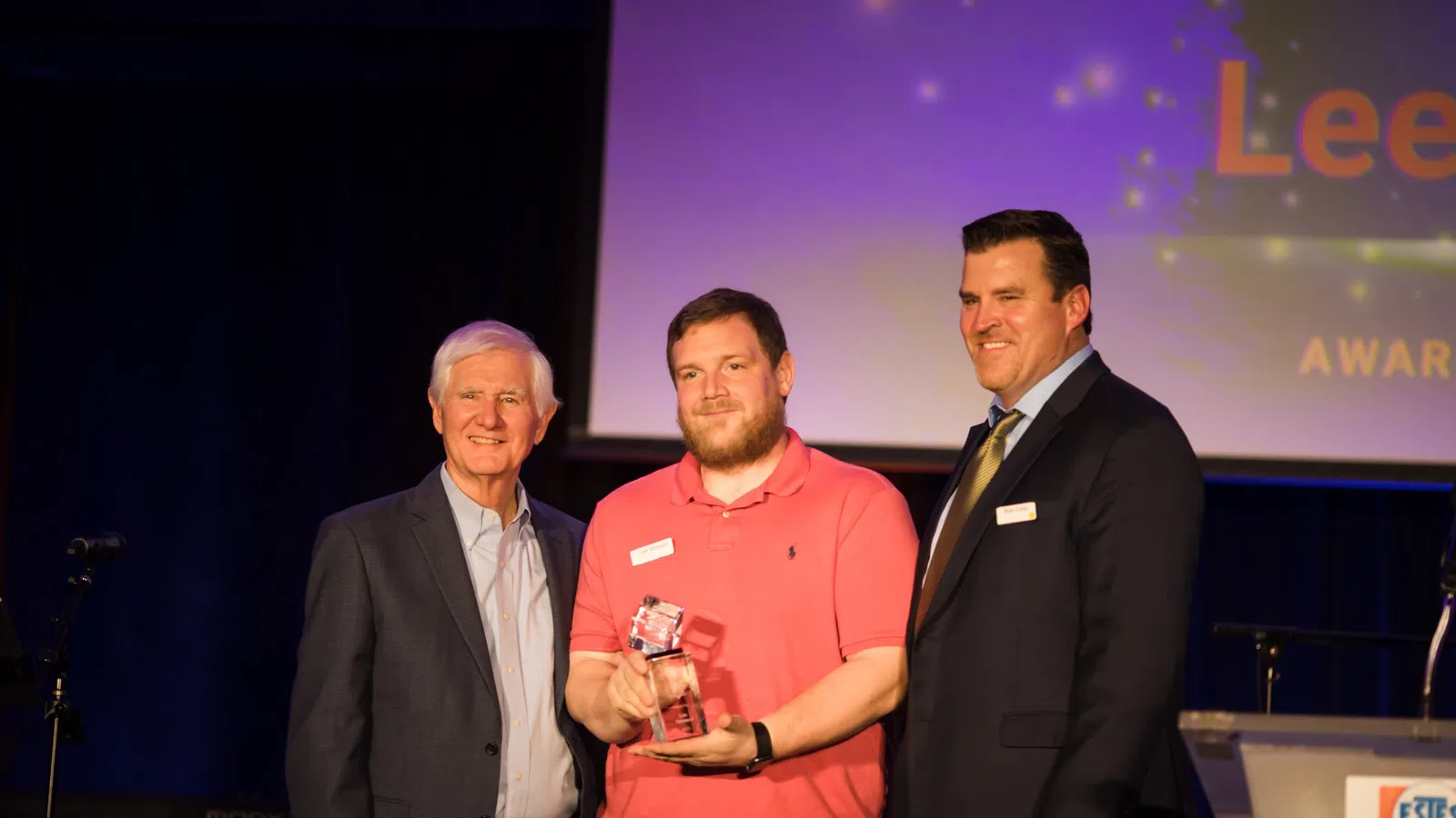 Three men smiling at an awards event, one holding a glass trophy, with overlaid text on screen behind them.