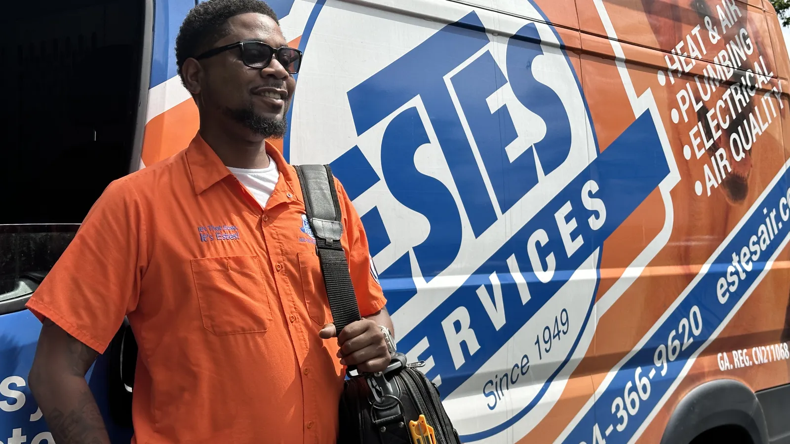 Estes Services Smiling service technician in orange shirt during summer time with tool bag stands beside Estes Services van with company branding
